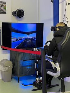 An adult sitting in a driving seat, wearing a VR headset and looking at a screen that shows the view from a car windscreen.